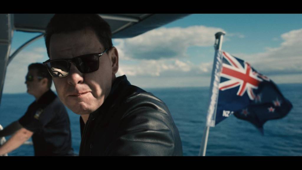 Still from the On P documentary showing Patrick Gower in sunglasses and slick black shirt on a boat with the New Zealand flag in the background